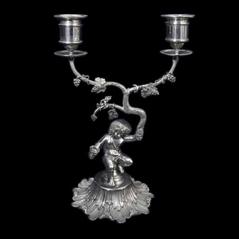 A pair of Bacchanalian 2 branch candelabra showing seated cherubs on circular bases holding wine cup in one hand and vine branches aloft in the other.The nozzles and drip pans decorated with fine bead borders.