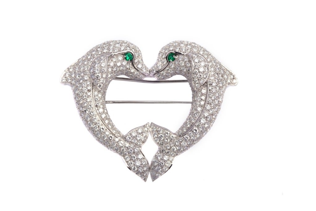 An elegant brooch manufactured by Cartier during the 1980s, representing a heart shape entailed by the encounter of two loveable dolphins. The item presents approximately 10cts of very fine quality brilliant cut diamonds and two emeralds (eyes) on a