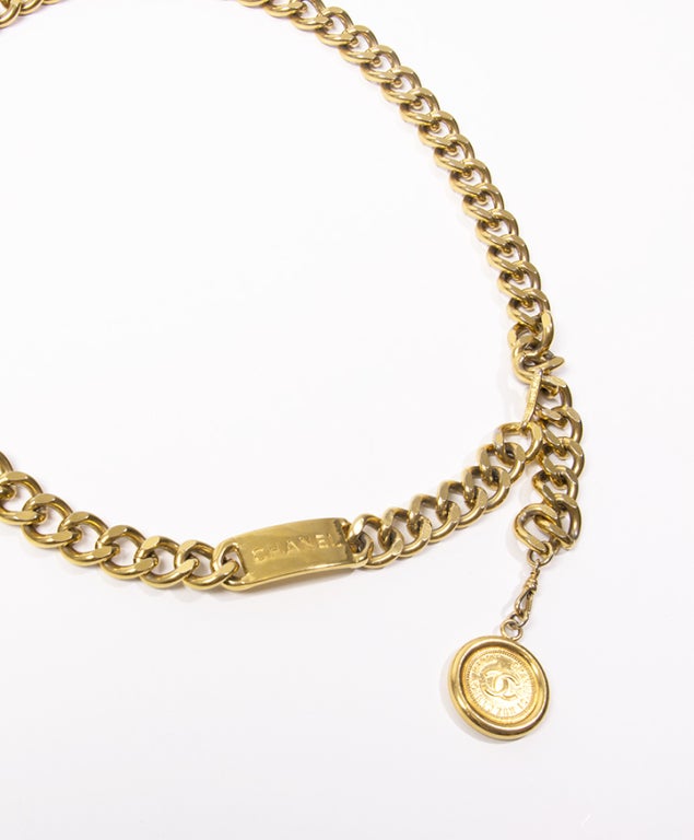 One-piece gold chain link belt with a logo plate and a round logo medallion on the end.