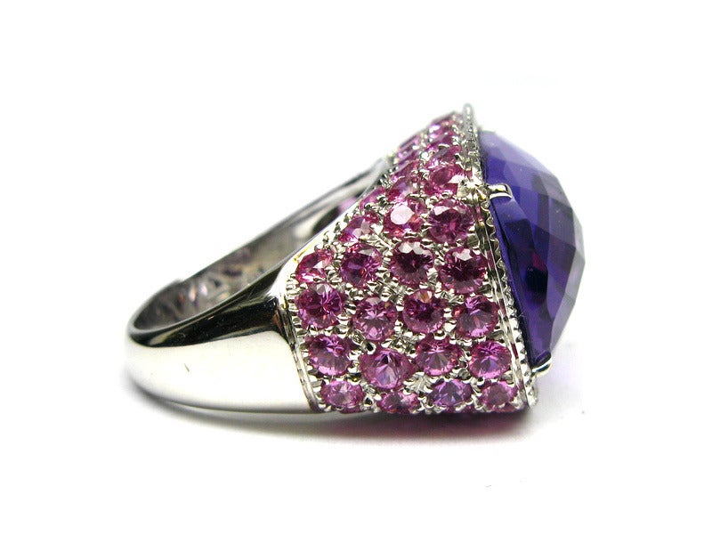 This gorgeous intricately detailed ring is 18kt white gold with a pave diamond halo and prong set pink sapphires along the sides. The center stone is a stunning checkerboard faceted cushion cut amethyst. This is the perfect statement ring for