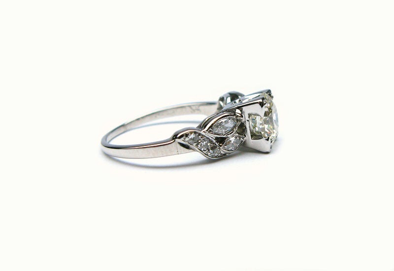 This unique and feminine Art Deco platinum engagement ring is a one of a kind piece. Featuring a 1.22 carat round diamond M color and SI1 clarity, the diamond is framed by chevron prongs that give it a squared look. Two marquise shape diamonds and a