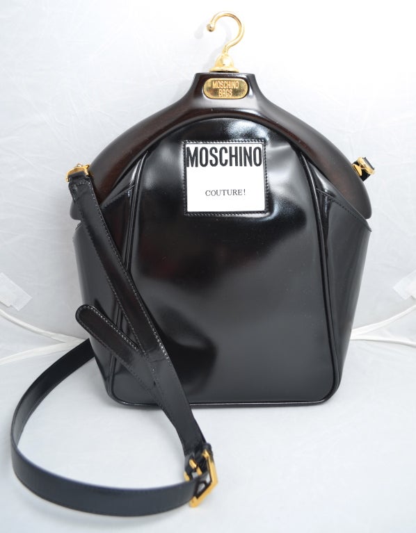 Franco Moschino lifetime coat hanger handbag with early Moschino Couture cloth label sewn into the front of the bag. Purse has a reddish brown wood coat hanger shaped top closure with gold details. There is a long adjustable shoulder strap with a