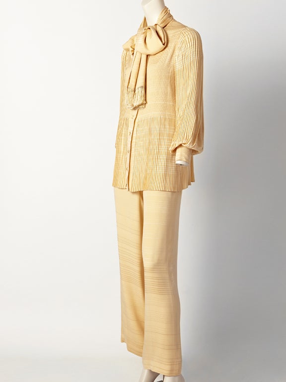 Andre Laug, peach tone, silk blouse and pant ensemble. Top has a generous tie with fringe,with full plisse sleeves. Pleating detail
continues beneath the bust and goes down to the hem. Trousers are high waist and wide legged.