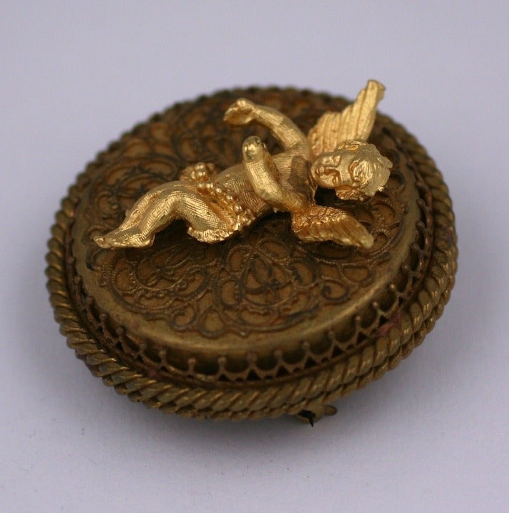 Charming brooch with applied cherub on a filigree base. There is a music box hidden beneath filigree cage. There is an attached pendant fitting as well. Self winding mechanism to operate music box. 2