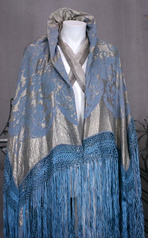 A dramatic art deco evening cape from the mid 1920s, France. Japanesque swallow tailed butterfly motifs in pale blue and silver lame broche. Knotted fringe of a darker blue silk and a criss cross lame ribbon interior closure.
Center back 52