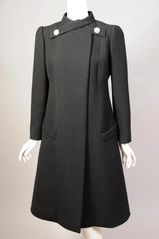 Pauline Trigere has designed an elegant evening coat in black wool with her signature diamante buttons. The collar has two tabs that cross and button. There is a series of concealed buttons as well. The two pockets have a wide band at the top which