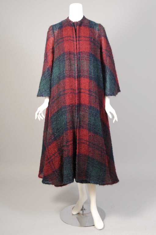 A feather light, unlined, mohair swing coat from Stavropoulos is designed in a deep red, green and navy blue plaid. The coat is collarless and it has no closures. The full bell sleeves echo the shape of the full coat. A large matching scarf is