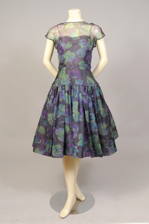 Rich purples, blues and greens are used for this stunning 1950's floral printed silk organza dress from Samuel Winston. The dress has a fitted bodice, a natural waistline, it is slim over the hips, with a full skirt. There is a zipper at the center