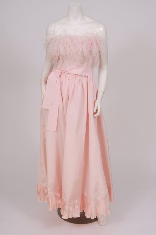 A pink silk faille strapless gown is accented with matching pink ostrich feathers at the bust line. A matching pink faille sash ties at the waist. The skirt is cut full with a pleated ruffle at the hemline. The dress has a center back zipper and it