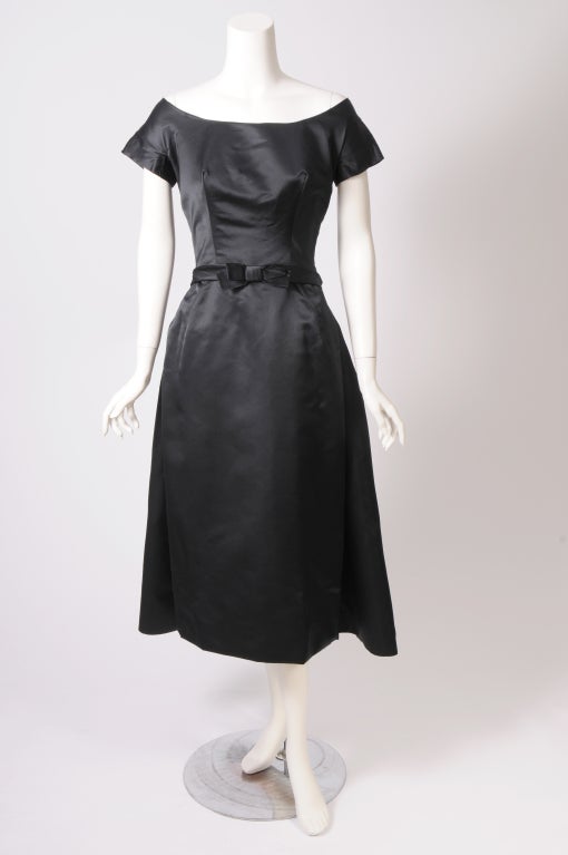 Inspired by the designs of Christian Dior this 1950's black silk cocktail dress is subtle and elegant. The scoop neckline sits off the shoulders with little cap sleeves. A flat bow accentuates the fitted waistline. It fastens at the front as part of