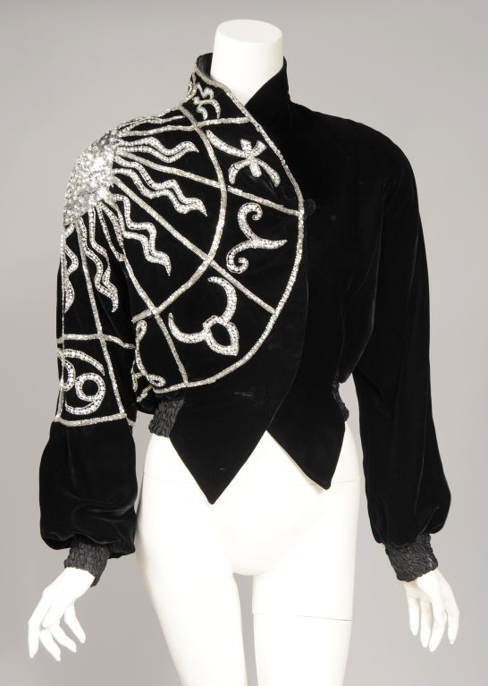 A striking beaded design is the focal point of this rich black velvet jacket from Bob Mackie. The signs of the zodiac surround a bold and graphic sun. They are worked in silver bugle beads, rhinestones and sequins. The cuffs and waistband are made