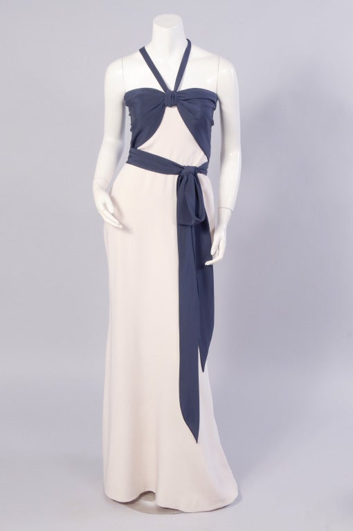 The bodice of this evening gown is draped with navy blue silk crepe over a strapless cream colored silk dress designed by Yves Saint Laurent for his Haute Couture line. The fitted bodice has a matching navy blue silk sash over a long and lean cream