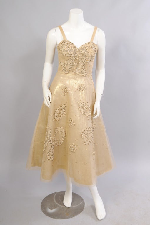 Glamorous gold lame is layered with golden tulle that is appliqued with large lace bows and bouquets of flowers. These appliques are beautifully beaded with pearls and gold sequins. The beads are also scattered on the tulle for added sparkle. The