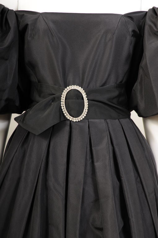 Pauline Trigere designed an elegant and chic evening gown from inky black silk taffeta. The low cut neckline allows the full sleeves to be worn off the shoulder.  The fitted bodice is acented by a matching wide black silk belt above the softly