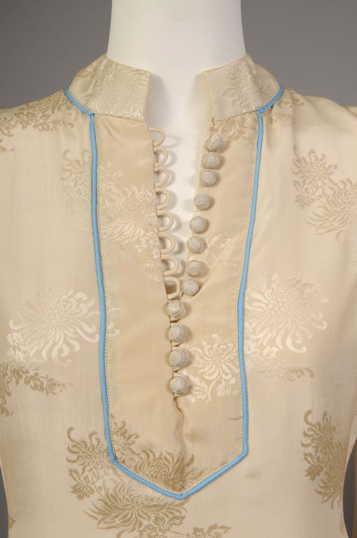 Tina Leser uses a woven China silk and Chinese design elements in this elegant cream silk dress. The tone on tone silk is woven with a pattern of chrysanthemums and trimmed with a narrow band of turquoise silk at the neckline. The dress has a