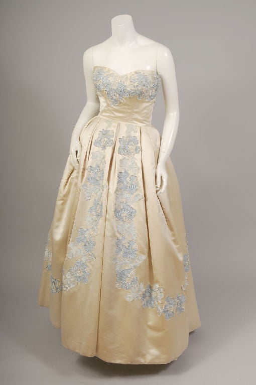 Luscious cream colored silk satin is embellished with tulle and silk ribbons in cream, blue and silver on the bodice and the full skirt of this elegant ballgown from Bergdorf Goodman.
The dress has a fitted strapless bodice above a full ball skirt.