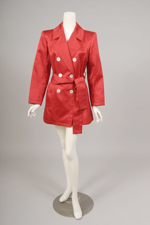 A classic trench from Yves Saint Laurent is given a bright new lease on life in brilliant red satin. The classic notched lapel, double breasted and belted style is accented with six mother of pearl buttons on the front. The jacket also has two