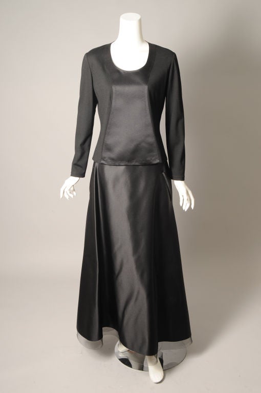Un llamativo vestido de noche de dos piezas de Geoffrey Beene realizado en una combinación de jersey de lana negro y satén negro. La parte central del top y toda la falda están hechas de satén. El resto del top es de punto. La falda está rematada