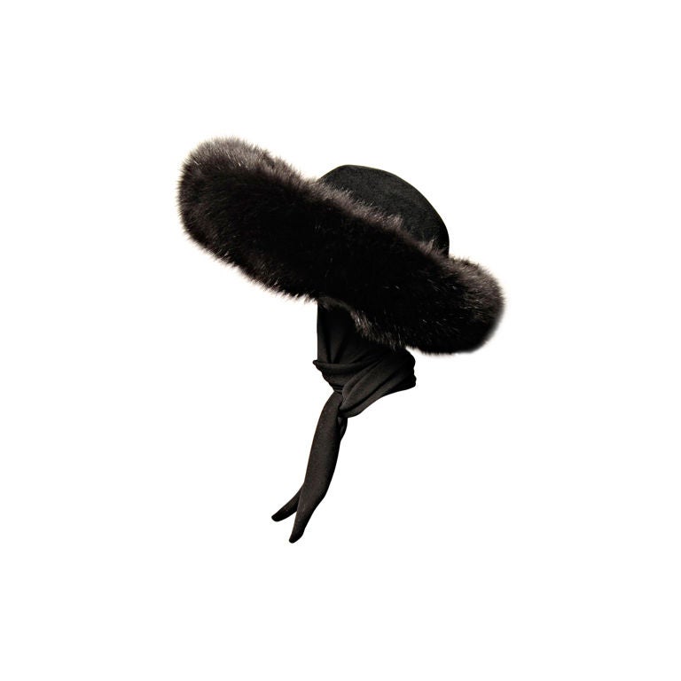 A deep band of luscious black fox surrounds the crown of this black wool felt hat, making it a glamourous choice for the upcoming winter months. For the coldest days the hat has an attached black jersey scarf to wrap and tie for added warmth. It is
