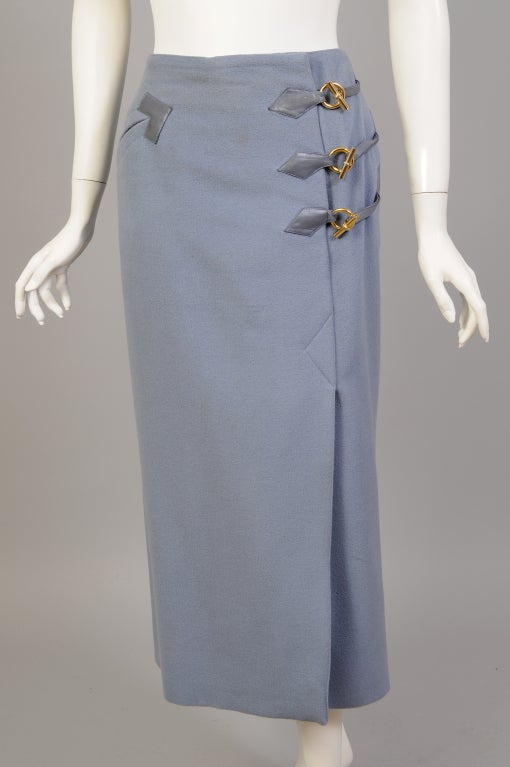 A blue wool skirt from Hermes is designed as a faux wrap skirt with blue lambskin and gold toned hardware as a decorative touch. The skirt is fully lined and there is a zipper concealed under the left side wrap. A diagonal pocket trimmed with