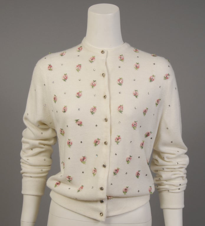 A creamy white wool sweater, Styled by Schiaparelli, is adorned with prong set rhinestones on the front, the back and the sleeves. In addition the front of the sweater has pink and green floral appliques with rhinestone centers. Prong set rhinestone