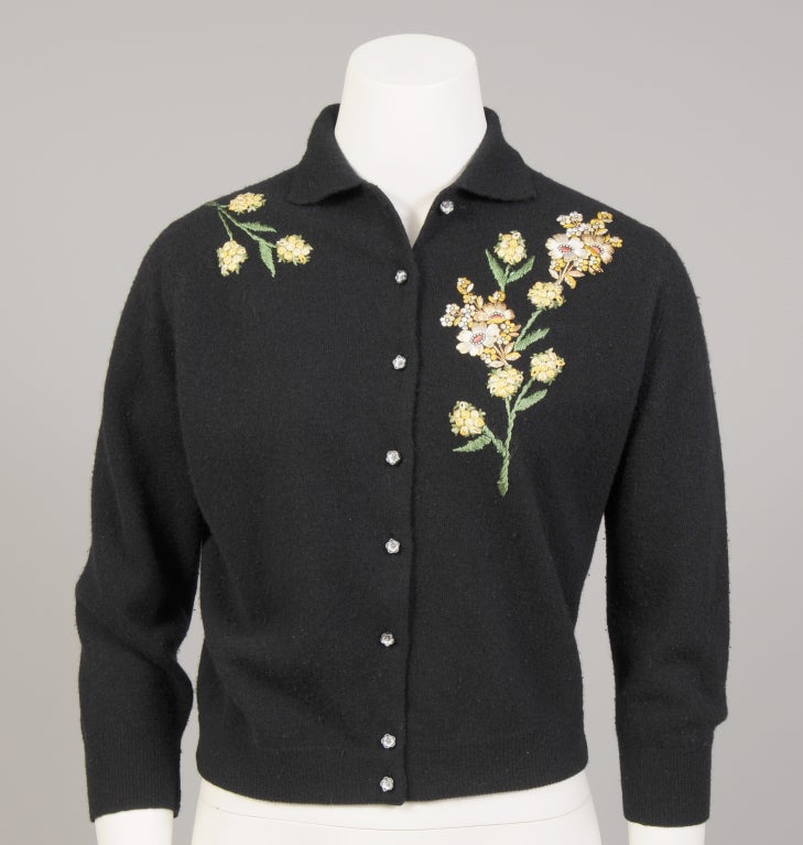 A black wool sweater, Styled by Schiaparelli, is decorated with a large floral motif on the right side and a smaller floral motif on the left. The sweater has a small collar and flower shaped buttons at the center front. It is in excellent