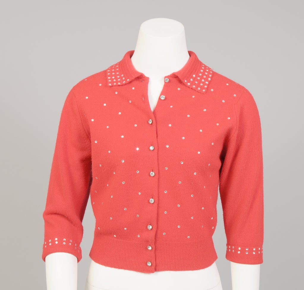 A bright red wool sweater, Styled by Schiaparelli in the 1950's, is embellished with rows of glittering rhinestones on the collar and cuffs. More rhinestones are scattered across the front of the cardigan sweater. Large prong set rhinestone buttons