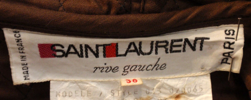 Si tratta di un cappotto trapuntato color kaki di Yves Saint Laurent, risalente alla fine degli anni Settanta. Il cappotto ha finiture in pelle marrone scuro, un cappuccio, due tasche a fessura sul davanti, chiusura frontale con alamari,  e una