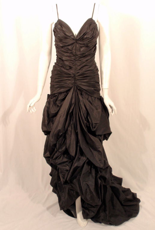 This is a glamorous long gown with a train. There is no designer name attached, but the gown was custom created. This gown has a ruched v-front fitted bodice with spaghetti straps and the skirt is poofed and draped, with a bustle like bow at the