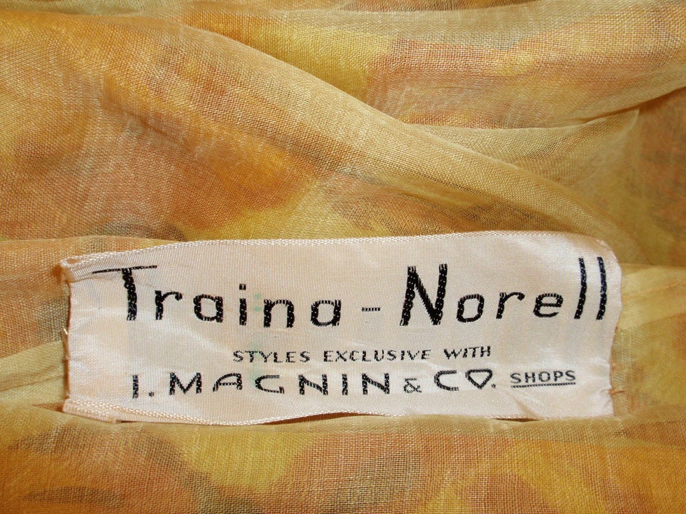 This is a charming cocktail dress from Traina-Norell. It is made of a yellow orange and brown floral print taffeta. It is sleeveless, with shoulder straps and a metal zipper on the back left side of the dress. The bow/sash is attached and wraprs