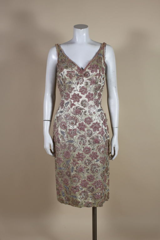 Stunning 1950's cocktail dress is done in a shimmering champagne brocade woven with a metallic floral pattern. Flowers are embroidered with textural faceted opaque pink and crystal clear glass beads. Fully lined with a back zipper.<br />
<br