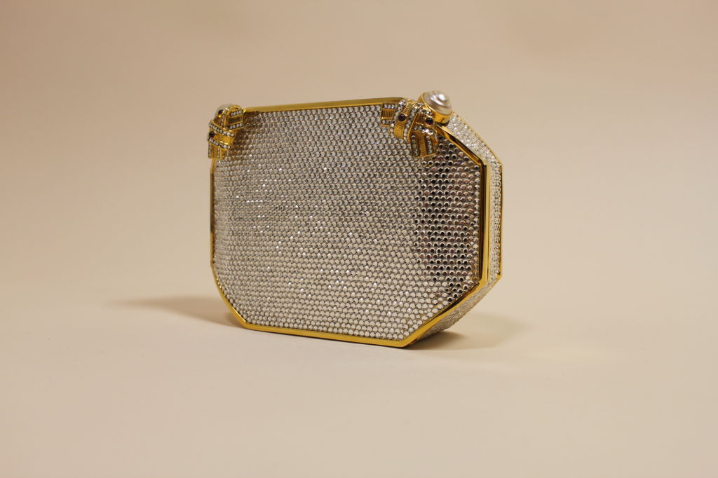 Elegant minaudiere from iconic designer Judith Leiber is done in an octagonal shape covered with sparkling crystal clear rhinestones on a luxe gold metal ground. Clutch snaps closed with molded ribbon shaped clasps decked with rhinestones and ruby