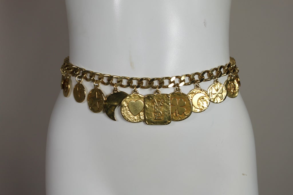 Fabulous gold-tone chain link belt from Yves Saint Laurent is decked with hammered gold coin charms that have various whimsical motifs such as clovers, hearts, butterflies and crescent moons.  Fastens with a textured gold