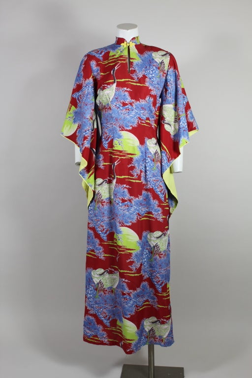 Beautiful 1940’s Hawaiian rayon dress features a vibrant crane print on a lipstick-red background. The print motif is that of cranes perched in sky blue branches set against lime green sunsets. Mandarin collar fastens with a pair of yellow frog