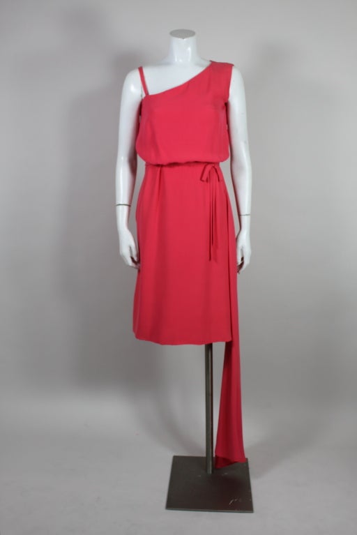 This 1960’s Mainbocher cocktail dress is made from a brilliant coral silk crepe. Cut asymmetrically, the dress has an attached side swag that cascades on the bias.The waist is accented by a delicate crepe bow. Dress is expertly finished with