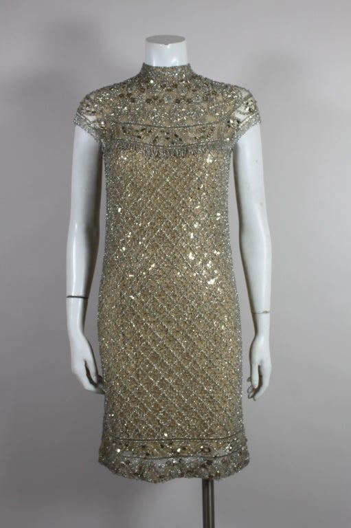 This fabulous 1960's cocktail dress features a heavily beaded overlay atop a nude lining. The dress shimmers with metallic silver beads and sequins elaborately sewn into a kaleidoscopic pattern. The neckline is fashioned into a trompe l'oeil collar