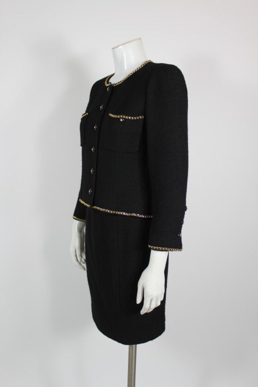 Iconic black wool bouclé tweed suit from Chanel. Trimmed in classic gold chain intertwined with black leather gold, this is a modern update on the classic Chanel ensemble. Fully lined in silk.

-14 buttons on jacket with one replacement button, 2