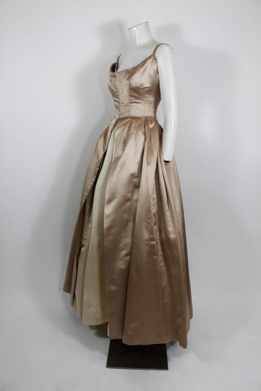 An elegant and lovely champagne satin ball gown from renowned American designer Nettie Rosenstein. An architectural and form-fitting bodice offsets a voluminous satin skirt with a peek-a-boo petal hem and overskirt. Partially lined with crinoline