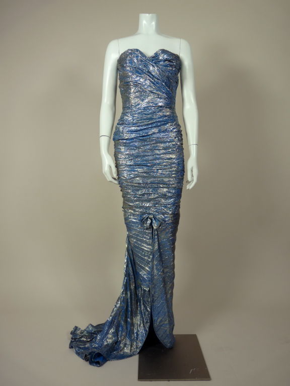 Dramatic blue and silver metallic brocade strapless gown from Emanuel Ungaro. Asymmetrically pleated bodice gathers to the side and cinches at the waist. Pleated skirt forms a modified bustle back that is accentuated by a matching rosette. Pleats