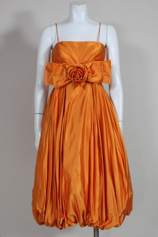 1950's tangerine hued silk satin cocktail dress has a voluminous bubble-hem skirt which is lined with an inner tulle petticoat. The center front of the pleated bodice is decorated with a self-double layer bow and sculpted rosette.  Back zip.<br