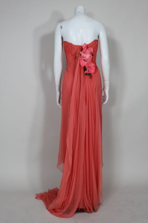 Diaphanous coral silk georgette evening gown from Irene features a hand pleated bodice and a cascade of silk flowers down the back. A flowing shoulder drape wraps around from flowers. Skirt is pleated from back waist and drops to a full-length hem.