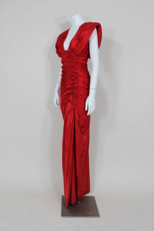 Stunning red silk jacquard gown from Arnold Scaasi has asymmetrical ruching at side front with pleats adorned by covered buttons. Bodice is structured with shoulder pads and features a plunging neckline with pleats from bust over shoulder to back