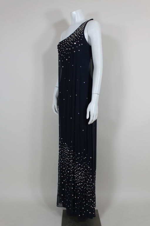 A stunning midnight blue unlabeled Halston one shoulder gown features a rhinestone encrusted chiffon overlay on a heavy matte jersey base. Jersey underdress clings to the figure while chiffon overlay flows freely. Glass rhinestones are prong set in