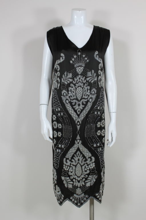 Ornately embroidered black satin 1920’s flapper dress features a fleur-de-lis inspired pattern made from hand sewn pearl white and clear glass beads. Hem is scalloped and edged with bands of beading. <br />
<br />
Measurements-<br />
Bust: 40”<br