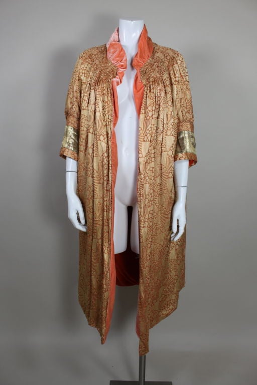 A gorgeous example of a 1920s opera coat, this piece is made from an apricot and gold silk lamé woven with an iconic geometric deco design. Modified kimono sleeves are decorated with a band of metallic gold lame. Neckline is hand smocked to create