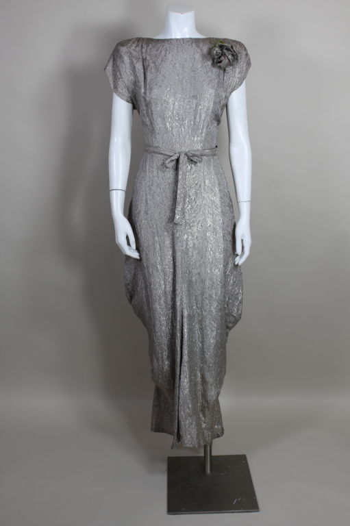 Fantastic silver silk lamé gown circa (1938-1942) has a structured shoulder silhouette with light padding and delicate pleating. Skirt is hand tacked at the sides in draping folds. Belt accentuates the waist. Detachable clusters of lamé rose