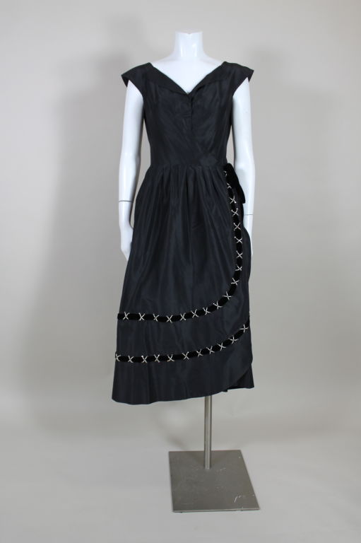 Fantastic fifties cocktail dress from Hattie Carnegie has a modern wrap closure that fastens with hook and eyes in the front. Skirt is pleated in the back for fullness and wraps with a scallop hem. Bands of velvet trim wrap around the hem accented