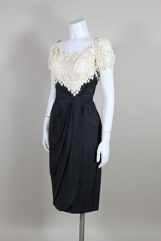 Fantastic 1950’s era cocktail dress from Ceil Chapman is made from black raw silk. Chapman’s signature pleating radiates from a side swag that falls into a sarong style. The bodice has a bust shelf that is decorated with ivory floral cotton lace