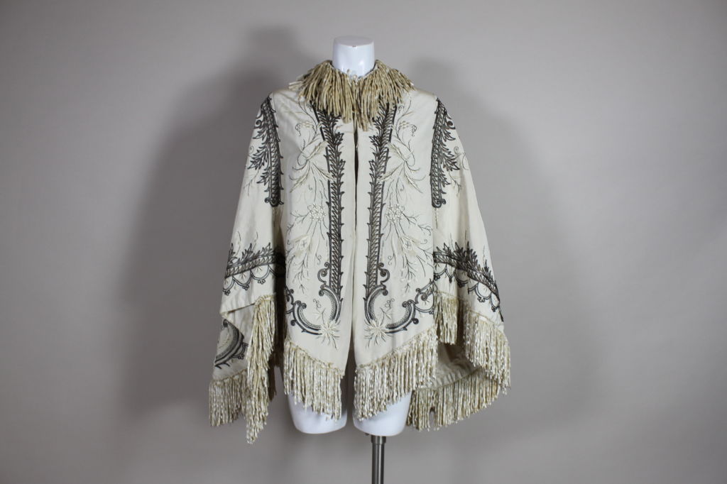 Fantastic Victorian cape is made from a cream silk twill with heavy embroidery. Cream and metallic bronze threads are stitched to create a scrolling floral motif. Cape is trimmed in cream silk chenille fringe. Fastens with hook and eye closure. <br