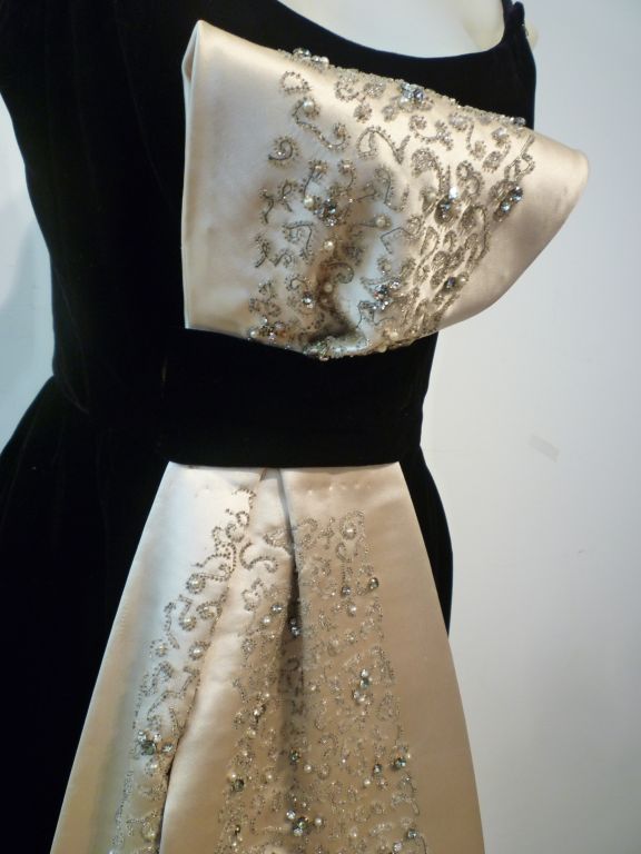 An exquisite 1960s couture black velvet column gown with a dramatic Asian-inspired beaded and embellished ivory satin obi bow. Heavily constructed bodice with boned underpinnings. An incredibly elegant and unique piece in a small size.  Approx US